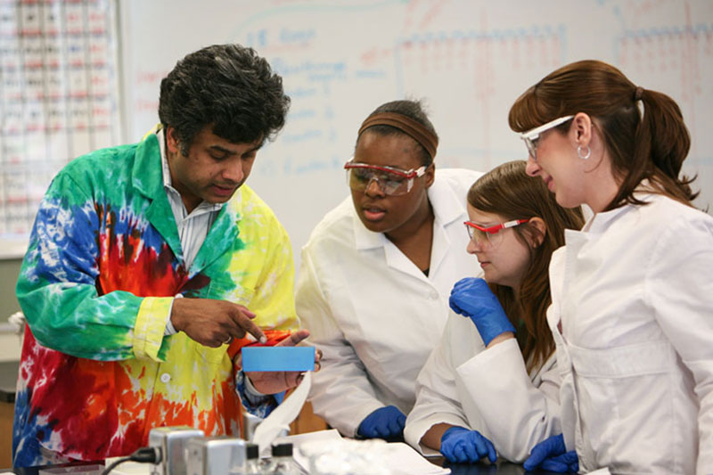 Faculty member in tie-dye lab coat reviews research with three students in white lab coats
